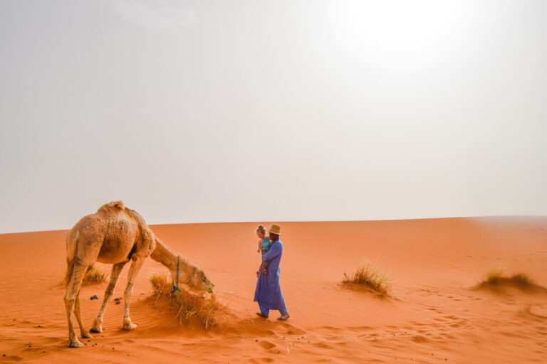 Moroccan man holds young child in the Sahara Desert as they both look at a camel standing next to them