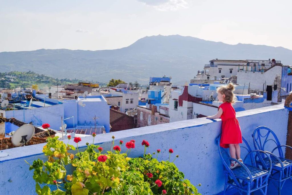 A young girl in a red dress, stands on a chair peering off the roof top in Chefchaouen.