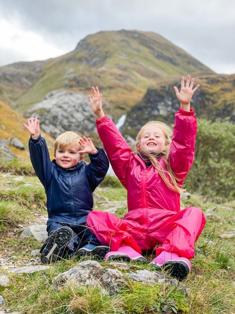 Young siblings sit on the grass with their arms in the air and huge grins on their faces.