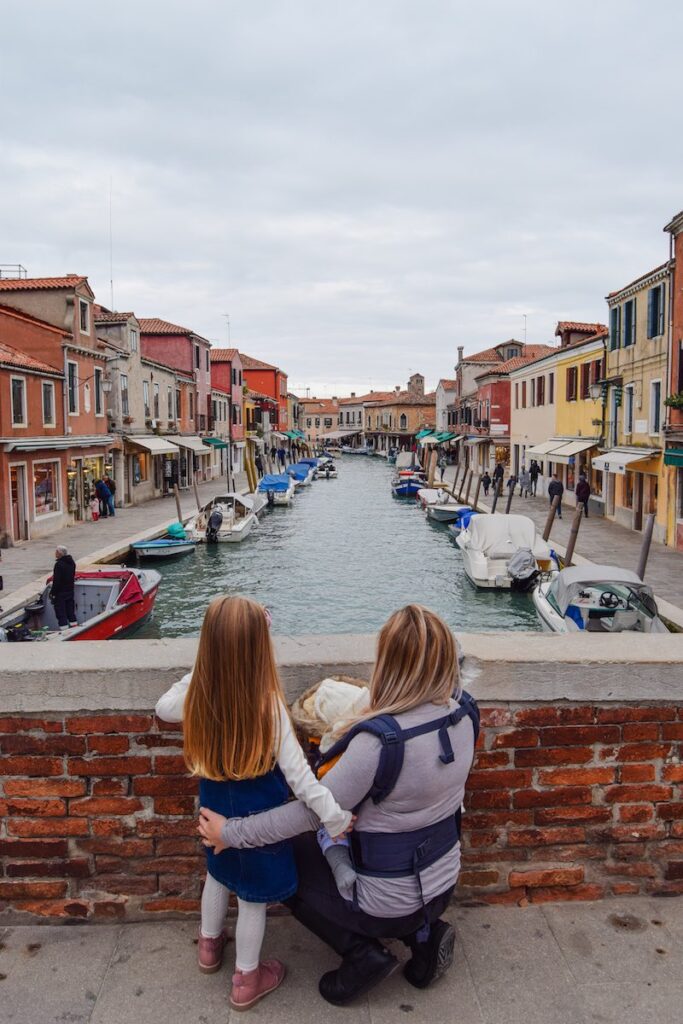 Mother and daughter standing together on a bridge in Murano, Venice, with colorful buildings in the background.