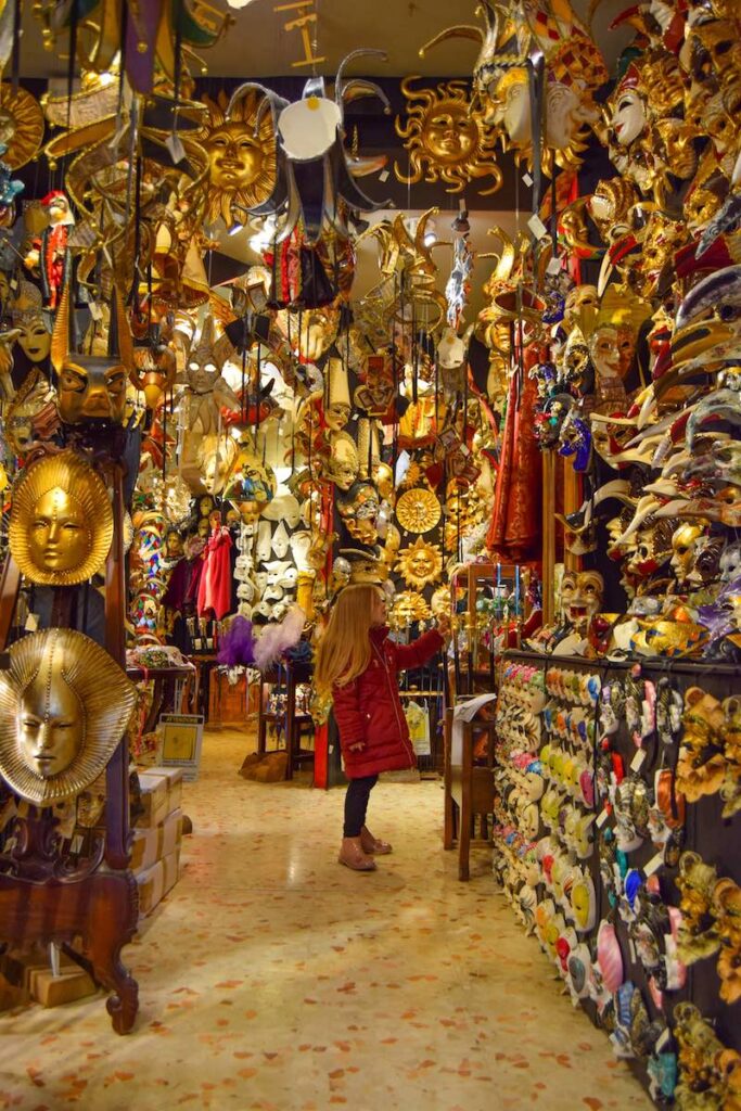 Young girl exploring a traditional Venetian mask shop filled with colorful handcrafted masks.