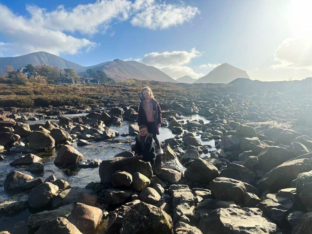 Two children playing and splashing in the River Sligachan beneath the historic Sligachan Old Bridge on the Isle of Skye, surrounded by mountains and lush greenery