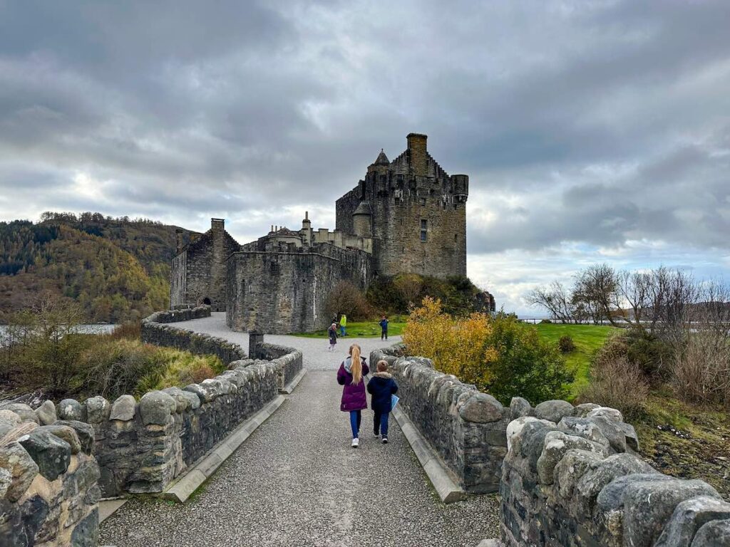 Two children walking along a path toward Eilean Donan Castle on the Scottish mainland, surrounded by scenic lochs and greenery